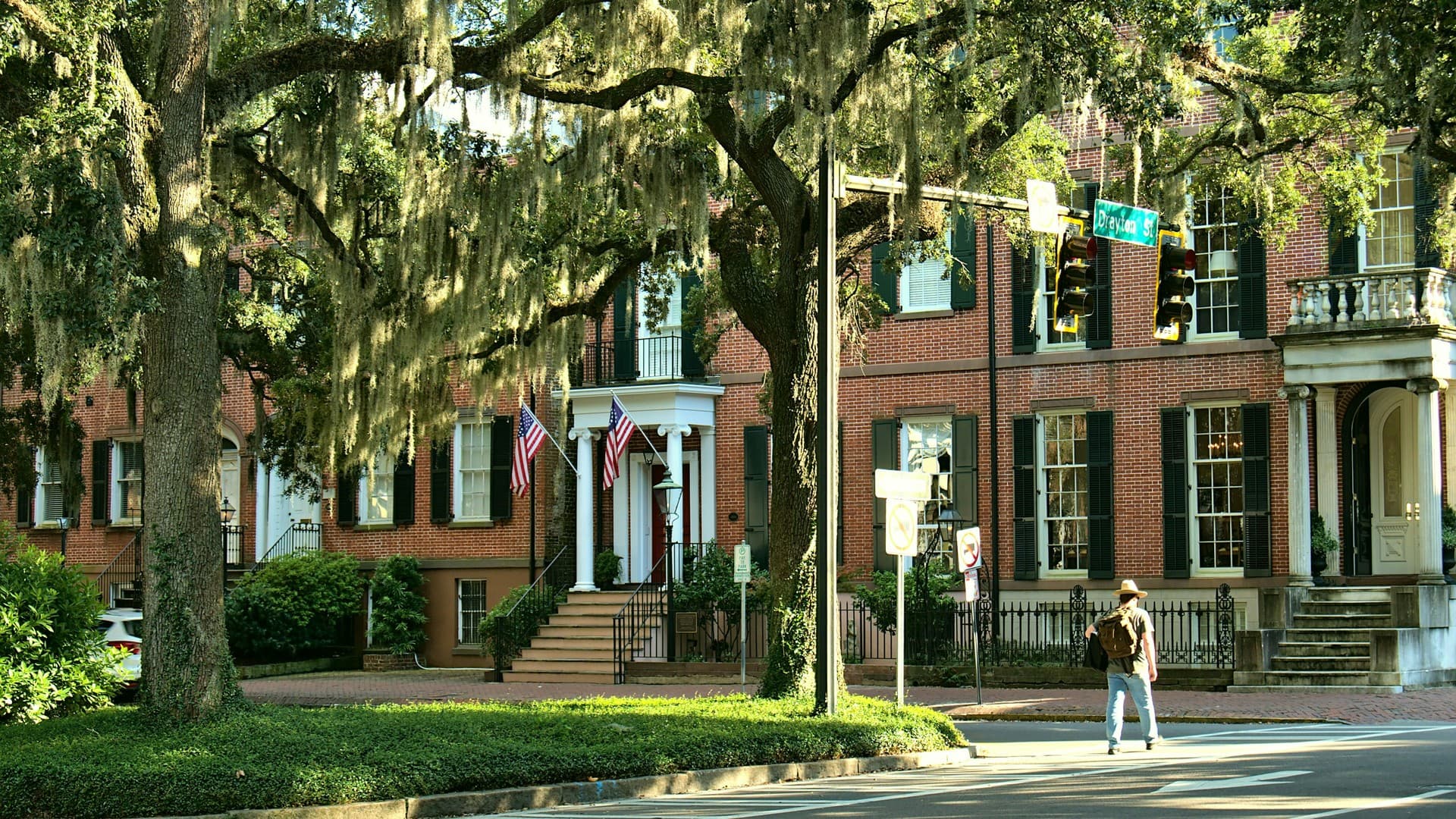 A shaded Savannah street corner under live oaks draped in Spanish moss, with historic brick townhouses and parking signage along the curb.